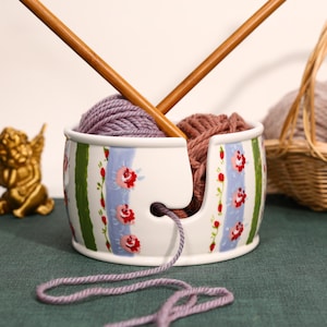 May include: A white ceramic yarn bowl featuring a vertical pattern of green stripes and light blue bands decorated with small red roses. The bowl contains two balls of yarn in purple and brown, with two wooden knitting needles crossed above.