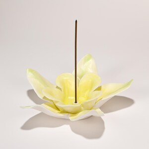 May include: A ceramic incense holder shaped like a flower with yellow petals and a white base. A brown incense stick is inserted in the center. The flower petals have a slight green tint at the tips.