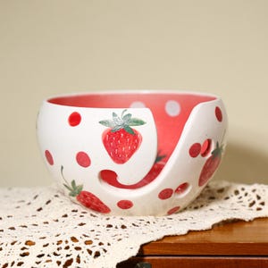 May include: A white ceramic yarn bowl featuring a hand-painted strawberry pattern and red polka dots. The interior is a solid red color, and the bowl includes a curved cutout slot for yarn to pass through, along with several small circular holes.