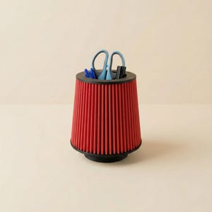 Peut inclure: Un filtre à air rouge transformé en pot à crayons. Le récipient cylindrique a une base et un bord noirs et contient des stylos bleus, un marqueur noir et des ciseaux bleus. Le fond est de couleur neutre.