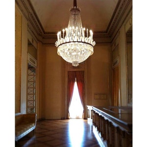 Peut inclure: Un grand lustre orné est suspendu dans une pièce à haut plafond. Le luminaire en cristal est éclairé, mettant en valeur les détails architecturaux de la pièce, notamment un parquet, des bordures dorées et des rideaux rouges. La pièce a un design classique et élégant.