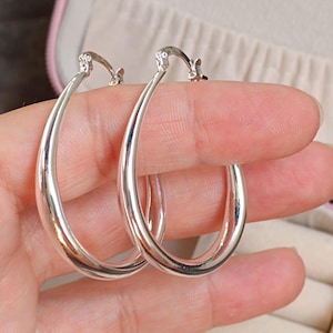 Puede incluir: Un par de pendientes de aro plateados pulidos. Los pendientes tienen forma ovalada y una superficie lisa y reflectante. Los pendientes están sostenidos por los dedos de una persona. Se muestran en una caja de joyería rosa.