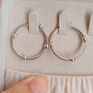 Può includere: Un paio di orecchini a cerchio argentati con un motivo a corda intrecciata. Ogni orecchino presenta tre piccole perle rotonde. Gli orecchini sono esposti in un astuccio rosa chiaro. Diametro: circa 3 cm.