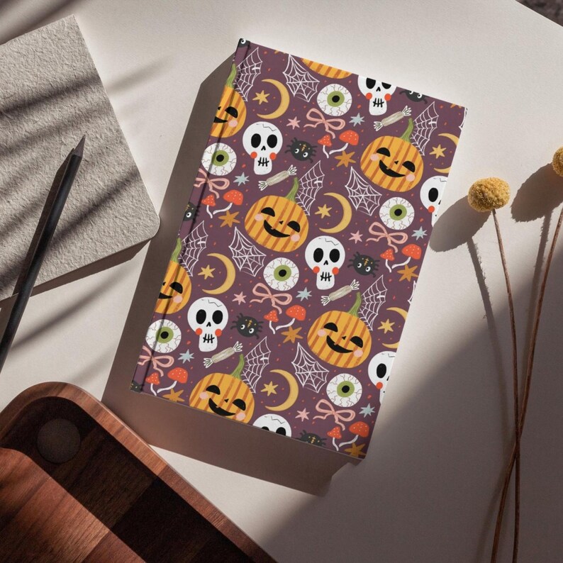 May include: A Halloween-themed journal with a purple cover featuring a repeating pattern of smiling jack-o'-lanterns, skulls, spiderwebs, eyeballs, and crescent moons. The journal is rectangular and appears to be closed, with a spiral binding.