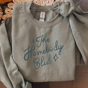 May include: Sage green sweatshirt with the text "The Homebody Club" in a teal script font. The sweatshirt is folded, with a knot tied at the top. The Gildan brand tag is visible.