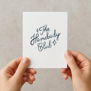 May include: A white card with the text "The Homebody Club" in dark teal script. The card is held by two hands against a neutral background. The text is surrounded by diamond shapes.