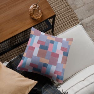 May include: Square decorative pillow with a colorful abstract geometric pattern in shades of pink, blue, brown, and white. The pillow is placed on a neutral-colored sofa, with a wooden table and glass vase in the background.