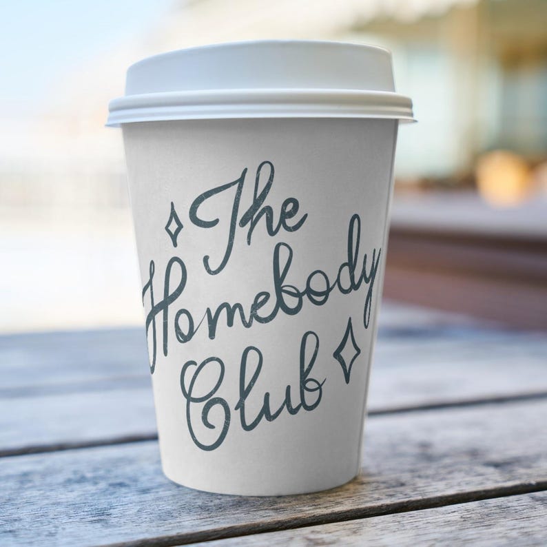 May include: A white paper coffee cup with a white lid. The cup has the text "The Homebody Club" in a dark teal script font, with diamond shapes on either side of the text. The cup is sitting on a wooden surface.