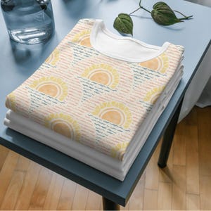 K&ouml;nnte beinhalten: Ein Stapel gefalteter wei&szlig;er T-Shirts mit einem gemusterten T-Shirt obenauf. Das gemusterte Shirt zeigt ein sich wiederholendes Design aus gelben Sonnen &uuml;ber blauen und rosa Wellenlinien auf cremefarbenem Hintergrund. Die Shirts liegen auf einem blauen Tisch.