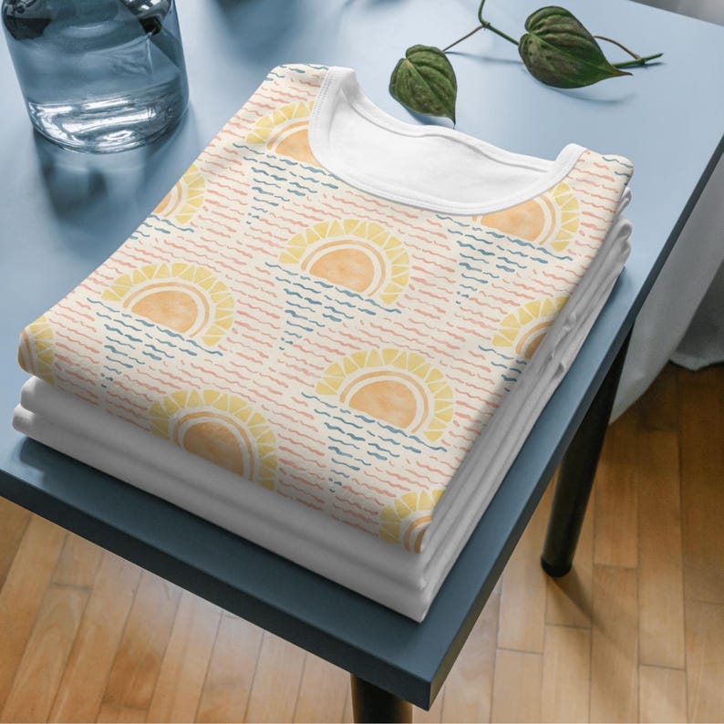 K&ouml;nnte beinhalten: Ein Stapel gefalteter wei&szlig;er T-Shirts mit einem gemusterten T-Shirt obenauf. Das gemusterte Shirt zeigt ein sich wiederholendes Design aus gelben Sonnen &uuml;ber blauen und rosa Wellenlinien auf cremefarbenem Hintergrund. Die Shirts liegen auf einem blauen Tisch.