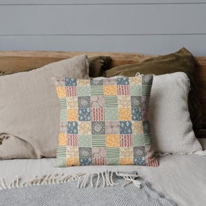 Może przedstawiać: Dekoracyjna poduszka z patchworkowym wzorem, przedstawiającym r&oacute;żne geometryczne kształty i wzory w odcieniach brązu, ż&oacute;łtego, niebieskiego i zielonego.