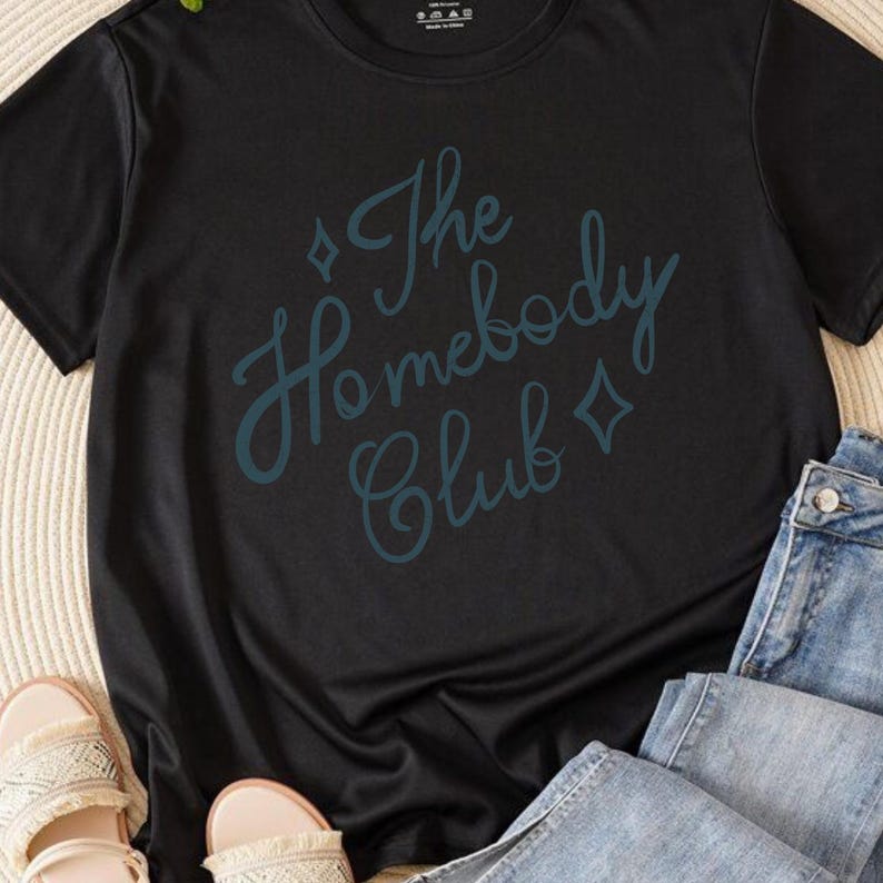 May include: Black t-shirt with the teal text "The Homebody Club" in a script font. The shirt is paired with blue jeans and beige sandals. The t-shirt is a casual wear item.