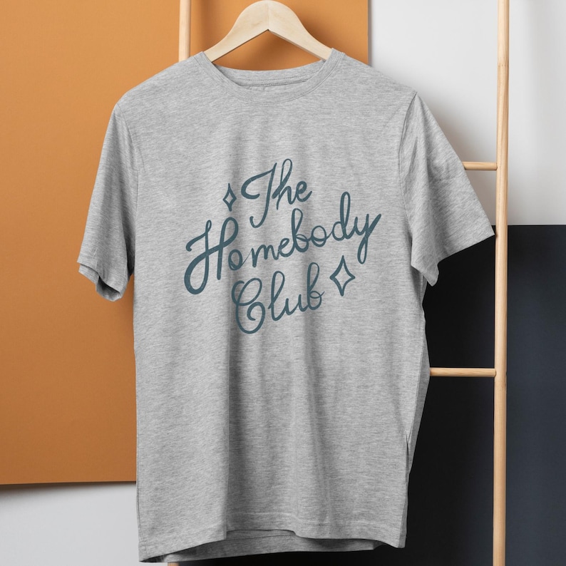 May include: A heather gray t-shirt with the text "The Homebody Club" in a teal script font. The design includes diamond shapes on either side of the text. The shirt is hanging on a wooden hanger.