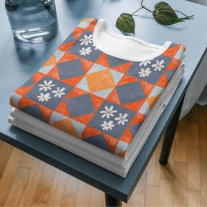 May include: Two white t-shirts, neatly stacked, with a patterned top layer. The top shirt has a geometric design in orange, grey, and light blue, with white daisy accents. The shirts are on a blue surface.