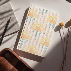 K&ouml;nnte beinhalten: Ein Hardcover-Notizbuch mit einem sich wiederholenden Muster aus gelben Sonnen &uuml;ber blauen und rosa Wellenlinien. Das Notizbuch liegt auf einer wei&szlig;en Oberfl&auml;che mit einem Bleistift, einem kleinen grauen Notizblock und einem Holztablett.