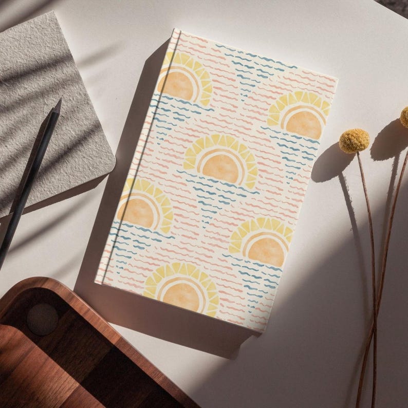 K&ouml;nnte beinhalten: Ein Hardcover-Notizbuch mit einem sich wiederholenden Muster aus gelben Sonnen &uuml;ber blauen und rosa Wellenlinien. Das Notizbuch liegt auf einer wei&szlig;en Oberfl&auml;che mit einem Bleistift, einem kleinen grauen Notizblock und einem Holztablett.