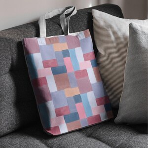 May include: A colorful tote bag with a geometric pattern of squares in shades of pink, blue, brown, and gray. The bag has gray handles and is resting on a gray sofa next to striped and solid pillows.