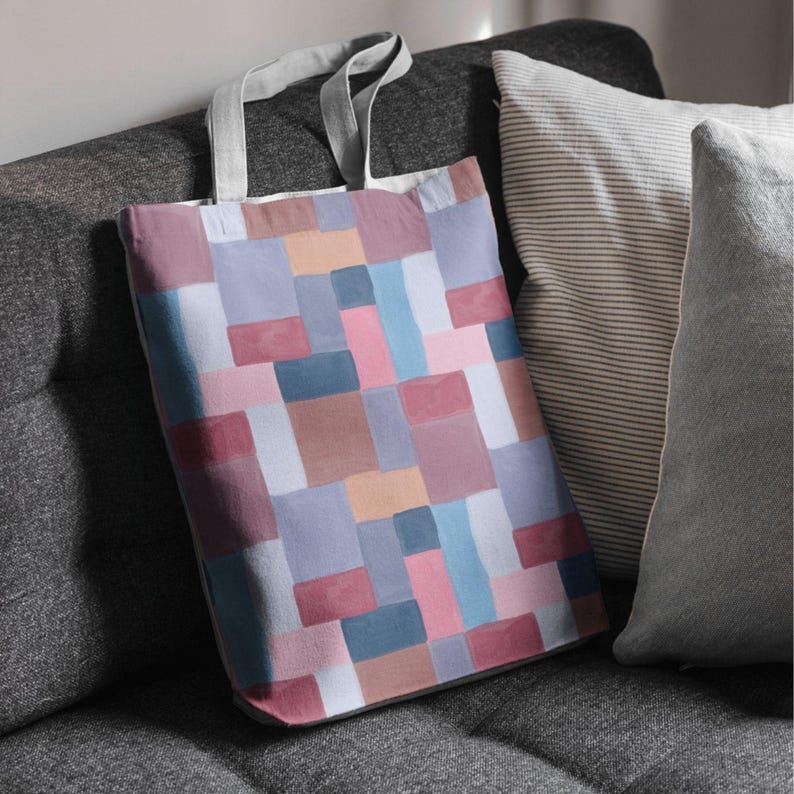 May include: A colorful tote bag with a geometric pattern of squares in shades of pink, blue, brown, and gray. The bag has gray handles and is resting on a gray sofa next to striped and solid pillows.