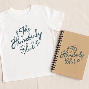 May include: White t-shirt with the text "The Homebody Club" in a dark teal script. A spiral-bound notebook with the same text is next to the shirt. The notebook has a brown cover.