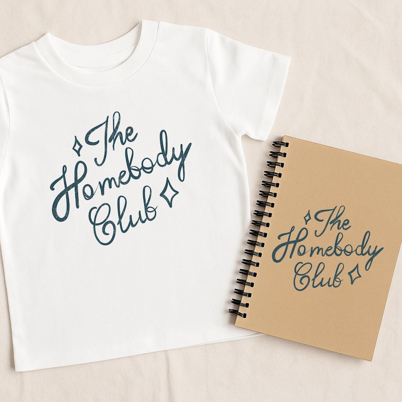 May include: White t-shirt with the text "The Homebody Club" in a dark teal script. A spiral-bound notebook with the same text is next to the shirt. The notebook has a brown cover.