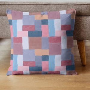 May include: Square throw pillow with a patchwork design. The pillow features a pattern of rectangular blocks in shades of pink, blue, gray, and peach. The pillow is resting on a wooden floor next to a beige sofa.