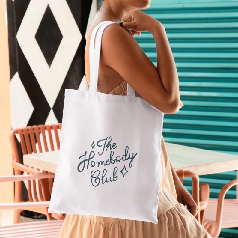 May include: A white tote bag with the text "The Homebody Club" in a script font. The bag has two handles and is being held by a person. The background includes a turquoise wall and a black and white geometric pattern.