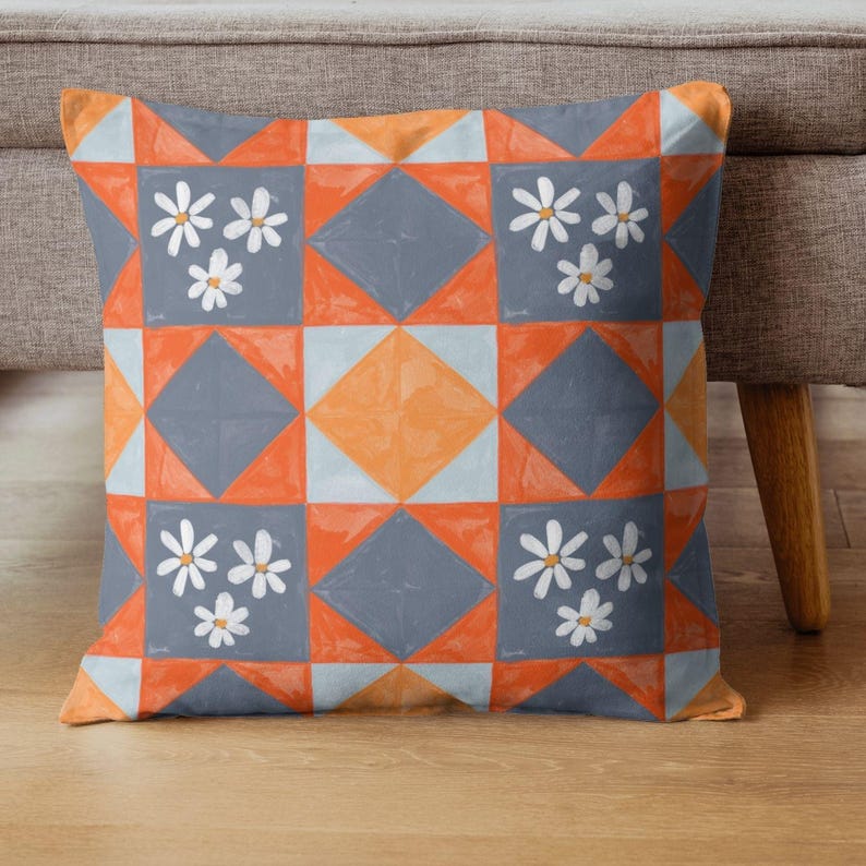 May include: Square cushion with a geometric pattern in orange, grey, and light blue. White daisy-like flowers are placed in the grey squares. The cushion is on a wooden floor next to a grey sofa.