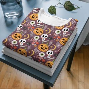 May include: A folded t-shirt with a Halloween-themed pattern featuring jack-o'-lanterns, skulls, spiderwebs, and eyeballs on a dark purple background. The shirt has a white collar and is stacked on top of other folded white shirts.