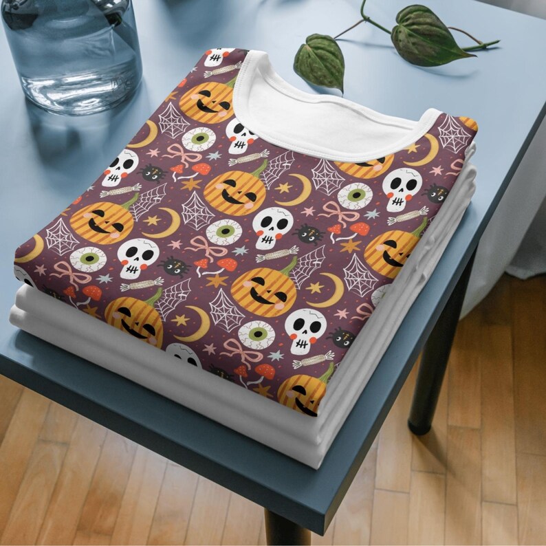 May include: A folded t-shirt with a Halloween-themed pattern featuring jack-o'-lanterns, skulls, spiderwebs, and eyeballs on a dark purple background. The shirt has a white collar and is stacked on top of other folded white shirts.