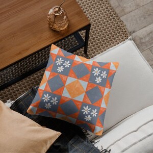 May include: Square cushion with a geometric pattern in orange, grey, and white. The design features diamond and square shapes with white daisy-like flower clusters. The cushion is on a light-coloured surface.