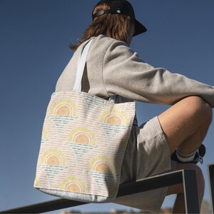 K&ouml;nnte beinhalten: Eine Canvas-Tragetasche mit einem sich wiederholenden Muster aus gelben Sonnen &uuml;ber blauen und rosa Wellenlinien. Die Tasche hat wei&szlig;e Tr&auml;ger und einen hellen Hintergrund. Das Design hat ein Strand- oder Sommerthema.