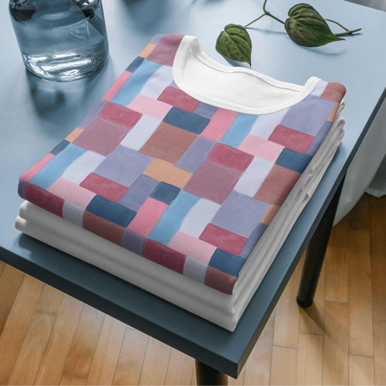 May include: A stack of folded t-shirts. The top shirt features a colorful geometric pattern in shades of pink, blue, brown, and purple. The shirts have a white crew neck collar. The shirts are stacked on a blue surface.