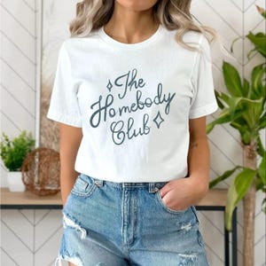 May include: White t-shirt with the text "The Homebody Club" in a teal script font. The design includes diamond shapes. The shirt is paired with denim shorts.