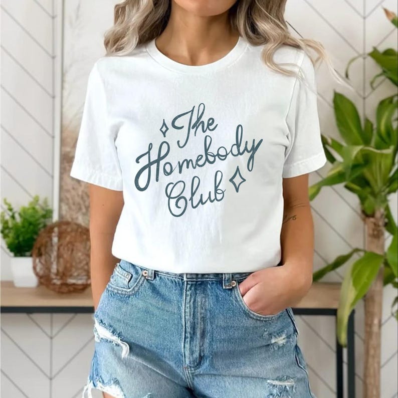May include: White t-shirt with the text "The Homebody Club" in a teal script font. The design includes diamond shapes. The shirt is paired with denim shorts.