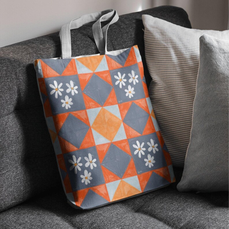 May include: A tote bag featuring a geometric design in orange, grey, and white daisy flowers. The bag has grey handles and is placed on a grey sofa next to striped and solid grey cushions.