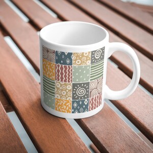 Może przedstawiać: Biały kubek ceramiczny z kolorowym wzorem geometrycznym. Wz&oacute;r składa się z kwadrat&oacute;w w r&oacute;żnych kolorach i wzorach, w tym spirale, kropki i linie. Kubek to świetny spos&oacute;b na dodanie odrobiny osobowości do porannej kawy lub herbaty.