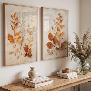 Two framed pressed leaf art prints hang on a white wall above a wooden console table styled with books, a small vase, a wooden bowl, and a glass vase with dried grasses.