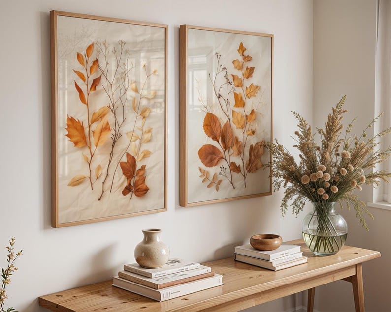 Two framed pressed leaf art prints hang on a white wall above a wooden console table styled with books, a small vase, a wooden bowl, and a glass vase with dried grasses.