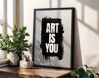 Puede incluir: Un cuadro enmarcado con las palabras "ART IS YOU" en blanco sobre un fondo negro con trazos de pincel. La impresión está en un marco negro y se encuentra sobre una superficie de madera, junto a una planta en maceta y un portalápices.