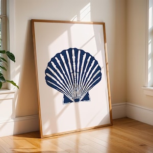 Puede incluir: Una impresión artística enmarcada con un diseño de concha marina azul marino sobre un fondo crema. La obra de arte está en un marco dorado y se apoya contra una pared. El diseño es una representación estilizada de una concha de vieira.