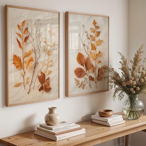 May include: Two rectangular wall art pieces in light wood frames, each containing pressed dried autumn leaves and thin branches on a cream-coloured background. The leaves display shades of orange, gold, and brown, arranged vertically.