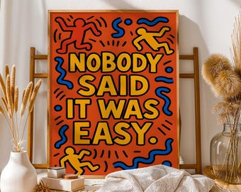 Puede incluir: Impresión artística naranja con el texto "NOBODY SAID IT WAS EASY" en amarillo y negro. La impresión presenta figuras rojas y amarillas y garabatos azules. La obra de arte está enmarcada en un marco de madera.