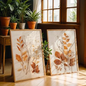 Two large framed pressed leaf art prints lean against a wooden floor in a sunny room with potted plants and wooden furniture.