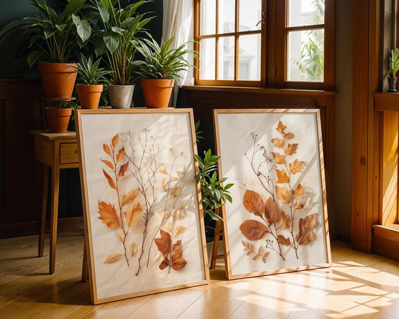 Two large framed pressed leaf art prints lean against a wooden floor in a sunny room with potted plants and wooden furniture.