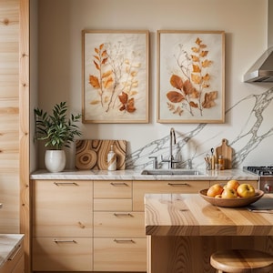The same two framed pressed leaf botanical prints are hung on a kitchen wall with marble backsplashes above wooden cabinets and a kitchen island holding a bowl of fruit.