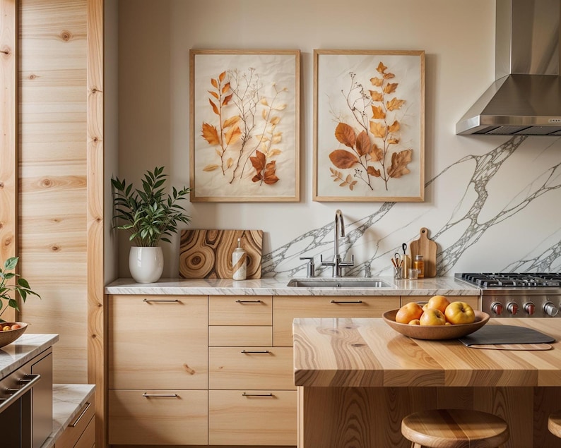The same two framed pressed leaf botanical prints are hung on a kitchen wall with marble backsplashes above wooden cabinets and a kitchen island holding a bowl of fruit.