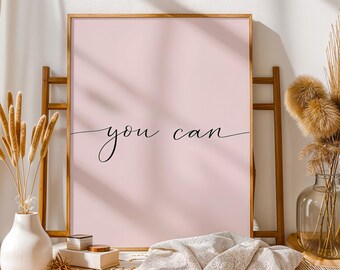 Puede incluir: Un estampado rosa enmarcado con las palabras "you can" en elegante escritura negra. La obra de arte se exhibe en un caballete de madera, con elementos decorativos como flores secas y un jarrón blanco.