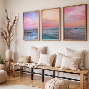 May include: A triptych of framed wall art prints depicting a beach at sunset with pink, orange, and blue hues. The artwork is displayed above a wooden bench with a black metal frame, featuring several beige throw pillows and a white blanket.