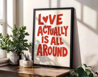 Puede incluir: Un estampado enmarcado con las palabras "LOVE ACTUALLY IS ALL AROUND" en letras rojas y en negrita. El estampado está en un marco negro y se encuentra sobre una superficie de madera, junto a una planta en maceta y un portalápices.