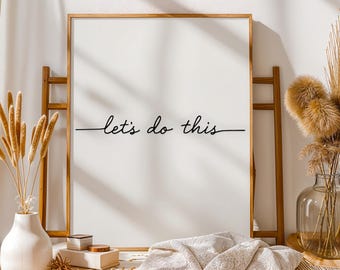 Puede incluir: Una impresión enmarcada blanca con las palabras "let's do this" en escritura negra. El marco es de color marrón claro. La impresión se exhibe con elementos decorativos, incluido un jarrón con plantas secas.